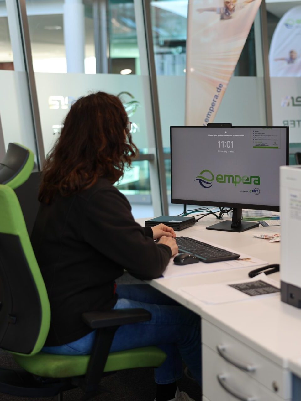 Mitarbeiterin, von hinten aufgenommen, sitzend an Schreibtisch in einem modernen Büro, umgeben von Computern und Büromöbeln